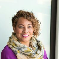 Smiling woman in a purple dress and scarf holding a tablet indoors.